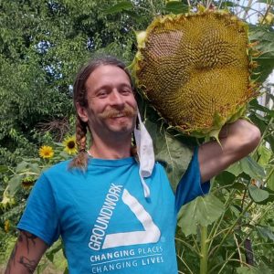 Photo of white man with semi long braided hair smiling and holding a giant sunflower